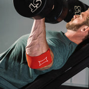 SLINGSHOT コンプレッションカフ2.0 レッドを着用しダンベルプレスを行う横方向視点。肘関節を支える位置とトレーニング中の安定感が伝わる描写