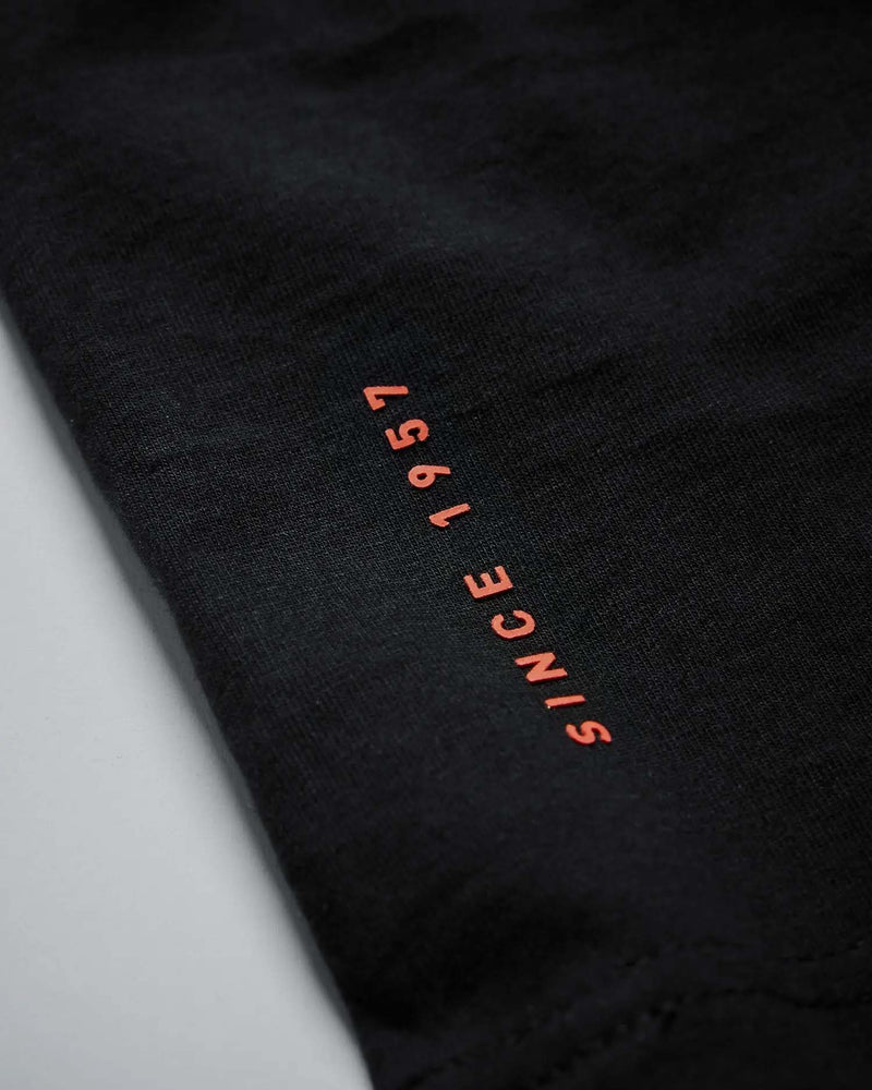 ELEIKO メンズ Tシャツ ブラックの生地と裾付近を斜めから拡大。黒い素材の質感と赤色の「SINCE 1957」プリント部分が分かる