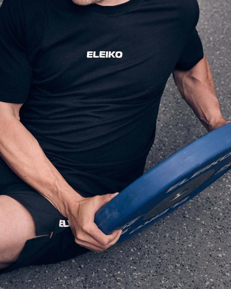 ELEIKO メンズ Tシャツ ブラックを着用してトレーニング器具を持つ男性を上方から捉え、胸元の白いロゴとフィット感が分かる