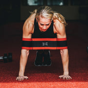 SLINGSHOT スリングショットプッシュアップ レッド＆ブラックを着用した女性が正面から腕立て伏せを行う視点。両上腕を支え、フォーム維持をサポートする構造。