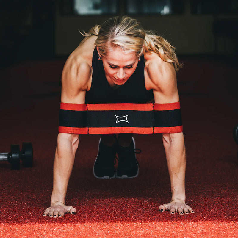 SLINGSHOT スリングショットプッシュアップ レッド＆ブラックを着用した女性が正面から腕立て伏せを行う視点。両上腕を支え、フォーム維持をサポートする構造。