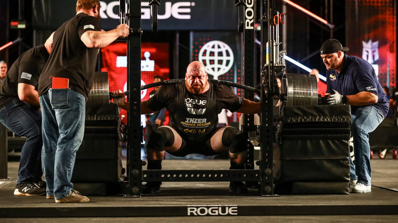 ROGUE CB-4 38mm キャンバーバー ブラック、パワーラック内でスクワットを行うアスリートを正面から捉えた競技シーン、曲線シャフトと大型プレート装着状態が確認できるスペシャリティバーベル