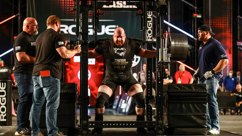 ROGUE CB-4 38mm キャンバーバー ブラック、スクワット中のアスリートを正面やや遠景から捉えた競技シーン、キャンバー形状シャフトと高重量プレート装着状態が分かるスペシャリティバーベル