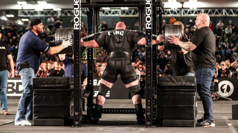 ROGUE CB-4 38mm キャンバーバー ブラック、スクワット中のアスリートを背面から捉えた競技シーン、曲線シャフトとプレート装着部、パワーラックとの位置関係が分かるスペシャリティバーベル