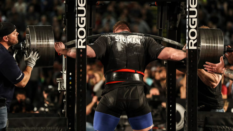 ROGUE CB-4 38mm キャンバーバー ブラック、パワーラック内でスクワット動作を行うアスリートを背面から捉えた競技シーン、湾曲シャフトと高重量プレート装着状態が分かるスペシャリティバーベル