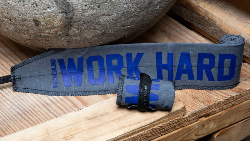 ROGUE ラップ グレーを木製ベンチ上で斜め上から捉えた構図、ブルーのWORK HARDプリントと巻いた本体・面ファスナーの厚みが確認できる配置